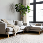 **

A bright, minimalist living room interior bathed in natural light. Soft grey walls with a jute rug under a light grey sofa. A chunky knit throw blanket is draped casually over the sofa. A fiddle-leaf fig tree stands in the corner near a window with sheer curtains, diffusing the morning light. A small, simple coffee table with a few carefully chosen objects (books, small plant) sits in front of the sofa.

**
